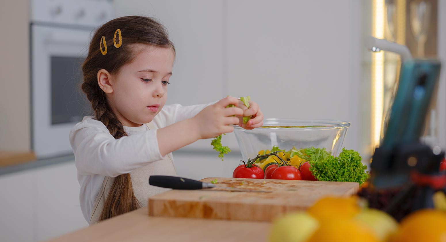 Incentivar hábitos saudáveis, Foto de uma menina na cozinha enquanto segura folhas de alface, a sua frente, está uma tábua de corte com tomate, alface e outras hortaliças e frutas