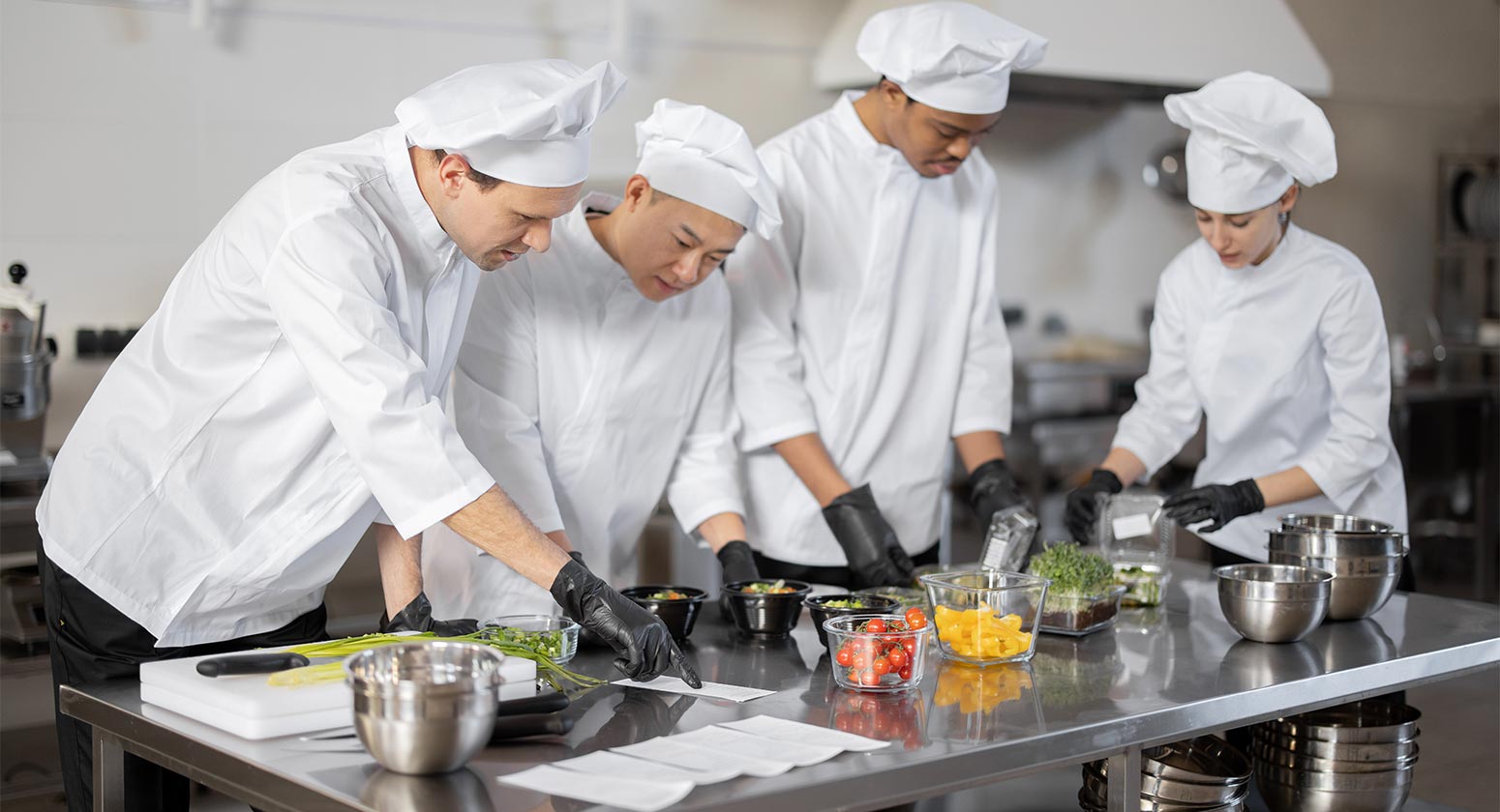 Contaminação Cruzada. Foto de cozinheiros em uma cozinha industrial enquanto preparam uma refeição.