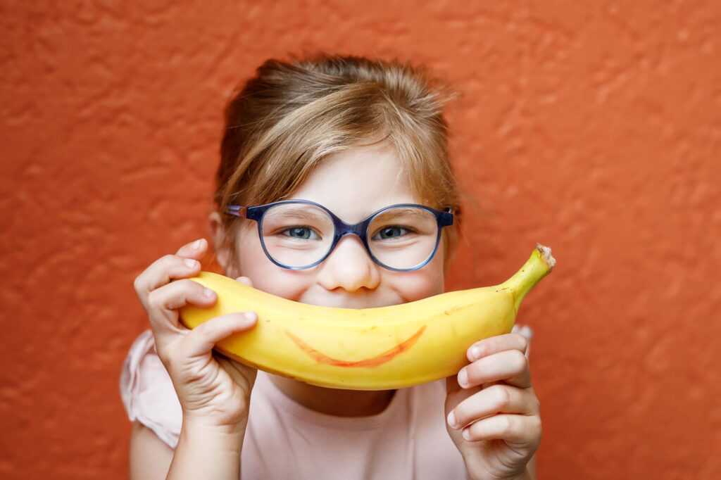 Imagem com uma menina sorrindo com o olhar enquanto segura uma banana onde ficaria sua boca.