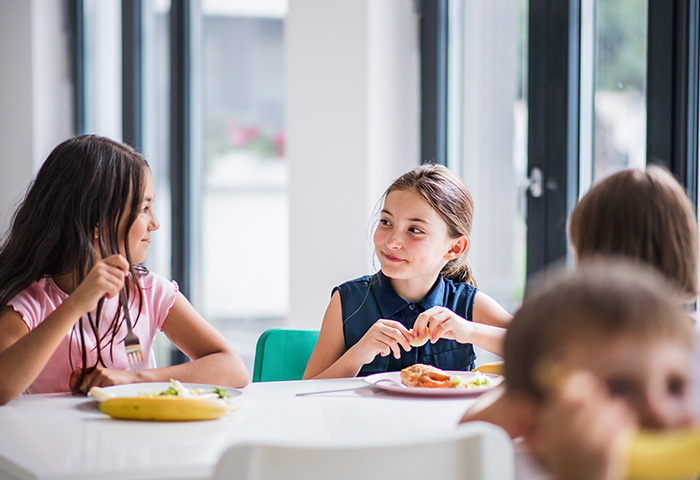 Duas meninas conversam enquanto almoçam juntas na escola. Legenda: A hora da refeição também é momento de socialização e aprendizado.