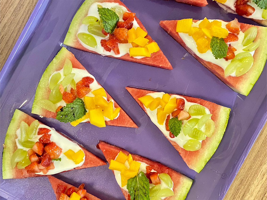 Fatias triangulares de melancia servidas como “pizzas” com cobertura de iogurte, uvas verdes, manga em cubos, morangos picados e folhas de hortelã, dispostas sobre uma bandeja roxa.