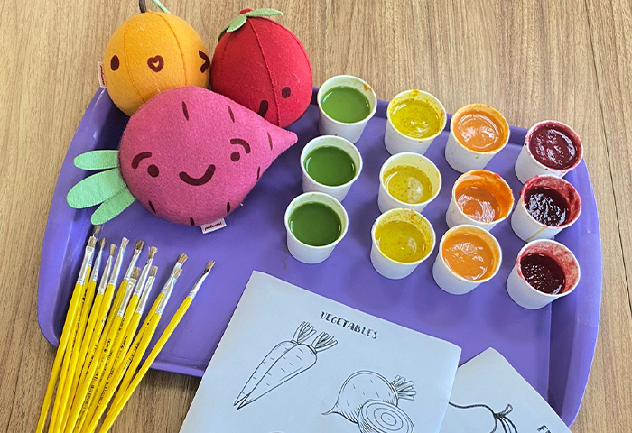 Materiais de atividade educativa com tema alimentar: pincéis, tintas coloridas em copinhos, pelúcias de frutas e vegetais, e folhas para colorir com ilustrações de legumes e frutas