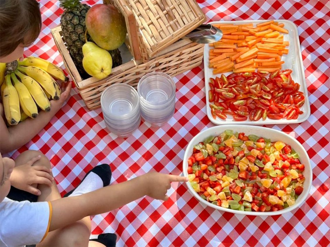 Crianças observam frutas frescas e legumes em uma oficina gastronômica escolar com mesa posta para lanche saudável.